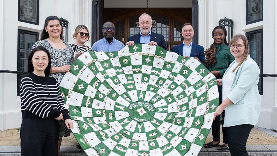 Loughborough University staff from the Vice-Chancellor's Office, Alumni Relations Team and Global Engagement Team holding a tapestry made by alumni in Abuja  in front of Hazlerigg Building on Loughborough University campus. They are all smiling in the sunshine and standing on the steps outside the doorway.